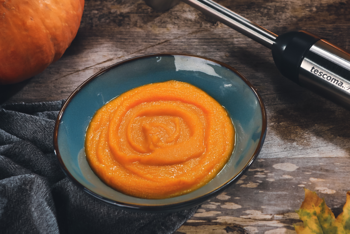 Gęste, pomarańczowe puree z dyni w ceramicznej, ciemnoniebieskiej miseczce z brązowym brzegiem. W prawym górnym rogu widać fragment blendera, a w lewym górnym rogu dynię.