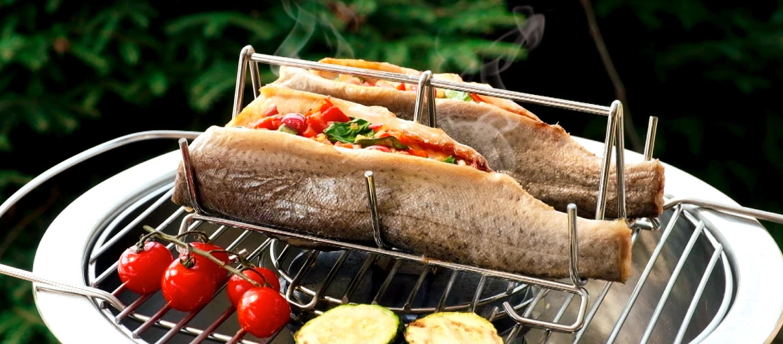 Opis: dwie faszerowane warzywami ryby w stalowym stojaku pieką się na grillu.
