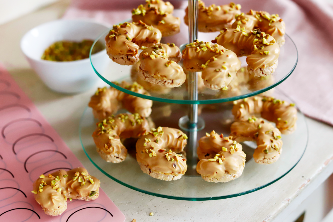 Rogaliki z polewą z czekolady karmelowej posypane pokruszonymi pistajami. Rogaliki zostały rozłożone na szklanej paterze. Obok patery leżą szablony do wypieków z narysowanymi kształtami.