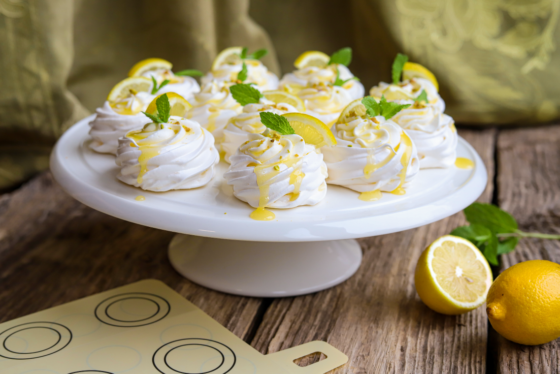 Mini bezy pavlovy z polewą cytronową i udekorowane listkami mięty leżą na białej porcelanowej tacy. Obok widać szablony do wypieków z narysowanymi kształtami.