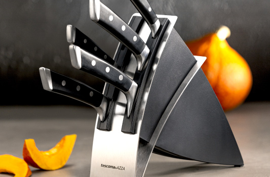 opis obrazka: masywny blok na noże w kolorze czarnym z dodatkiem stali, w środku pięć stalowych noży o wyprofilowanych, czarnych rękojeściach z nitami. W tle pomarańczowa dynia, jedna cała oraz kawałki pokrojonej dyni.