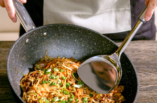 opis obrazka: metalowa łopatka trzymana przez osobę, w tle azjatyckie danie z makaronem, warzywami i mięsem usmażone w patelni wok