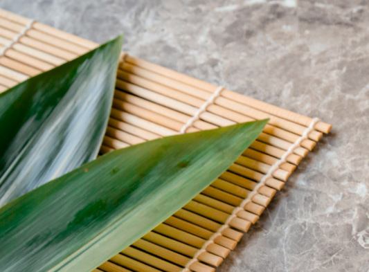 opis obrazka: na kamiennym blacie leży bambusowa mata do zwijania sushi, a na nim dwa duże ciemnozielone liście bambusa.