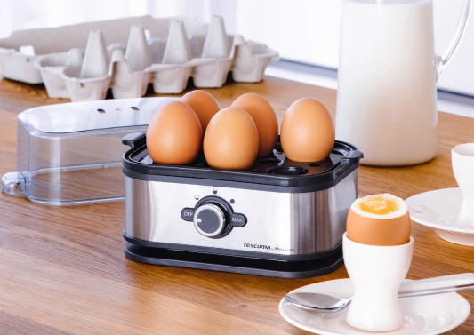 Na drewnianym blacie stoi srebrno-czarny elektryczny jajowar, a w nim pięć jajek. Szóste jajko znajduje się na pierwszym planie. Jest ugotowane na miękko, znajduje się w białym kieliszku, który stoi na białym spodku z metalową łyżeczką. Za jajowarem stoi dzbanek z mlekiem, pokrywa jajowaru oraz puste tekturowe pudełko po jajkach.