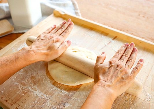 Wyprostowane dłonie wałkują ciasto na pizzę przy pomocy prostego drewnianego wałka. Ciasto leży na drewnianej stolnicy oprószonej mąką.
