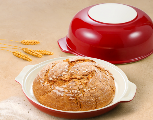 Na jasnej powierzchni leży otwarta ceramiczna okrągła forma na chleb z czerwoną, błyszczącą glazurą. Na pierwszym planie znajduje się jej płytki spód, a w nim leży okrągły, apetyczny, upieczony, lekko popękany i oprószony mąką bochenek chleba. Po lewej stronie widać cztery źdźbła pszenicy.
