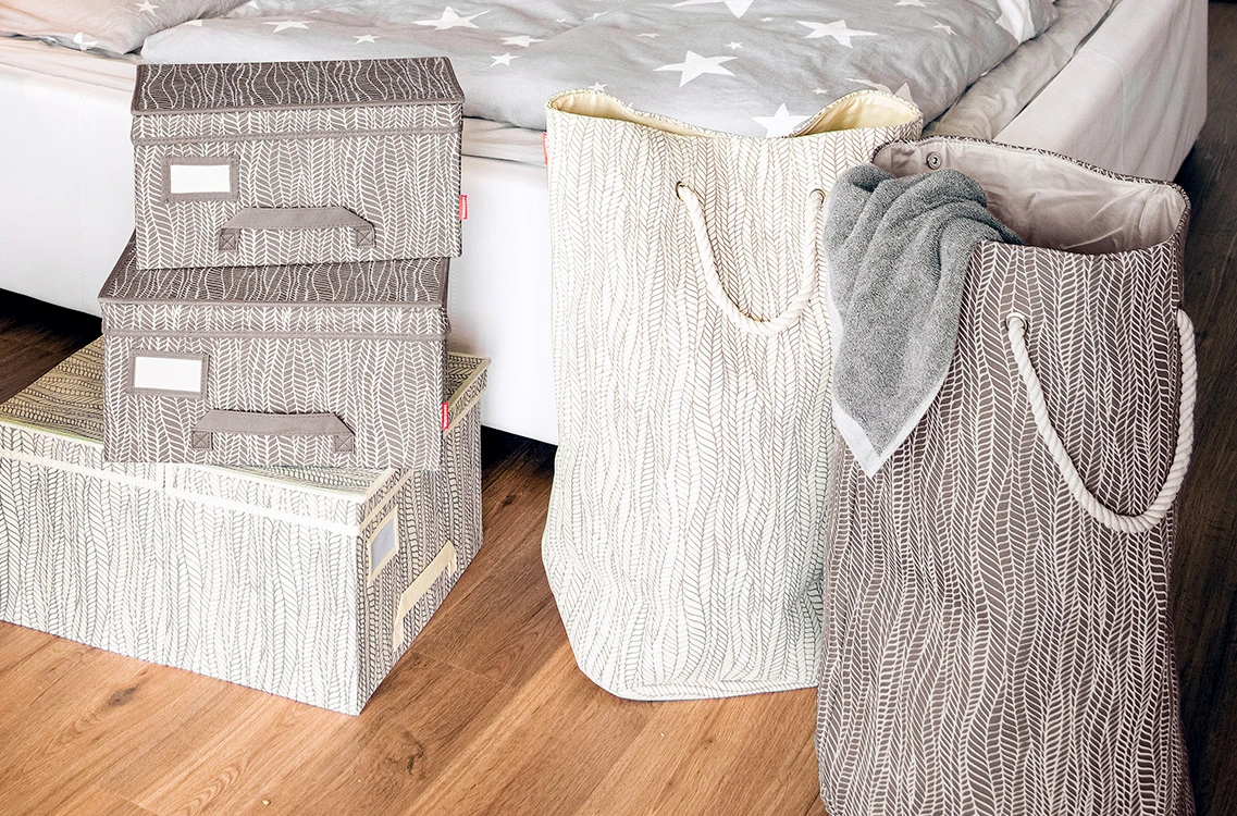 Widać kawałek sypialni - na drewnianych panelach stoi posłane łóżko z pościelą w jasnych kolorach. Przed łóżkiem na podłodze stoją pudełka na odzież w jasnobrązowym i kremowym kolorze, a po prawej dwie duże torby na pranie w tych samych kolorach, co pudełka. Torby mają sznurkowe uchwyty, a z brązowej torby wystaje szary ręcznik.