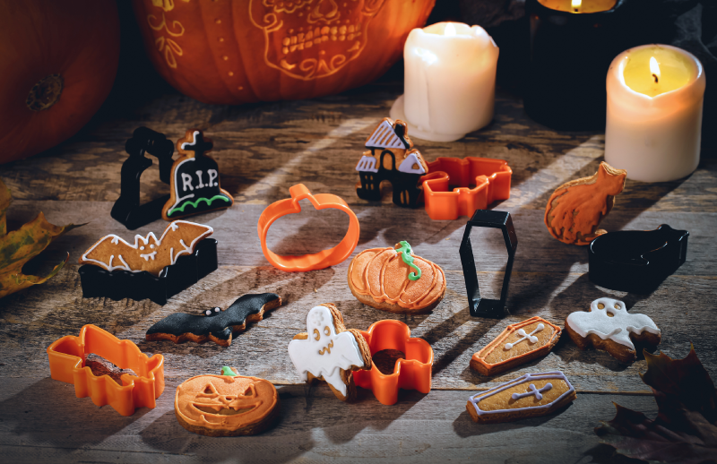 Na drewnianym stole leżą foremki i pokryte koloworym lukrem ciasteczka w halloweenowych kształtach: dyni, grobu, kota, nawiedzonego domu, ducha, nietoperza i trumny. W prawym górnym rogu stoi zapalona świeca.