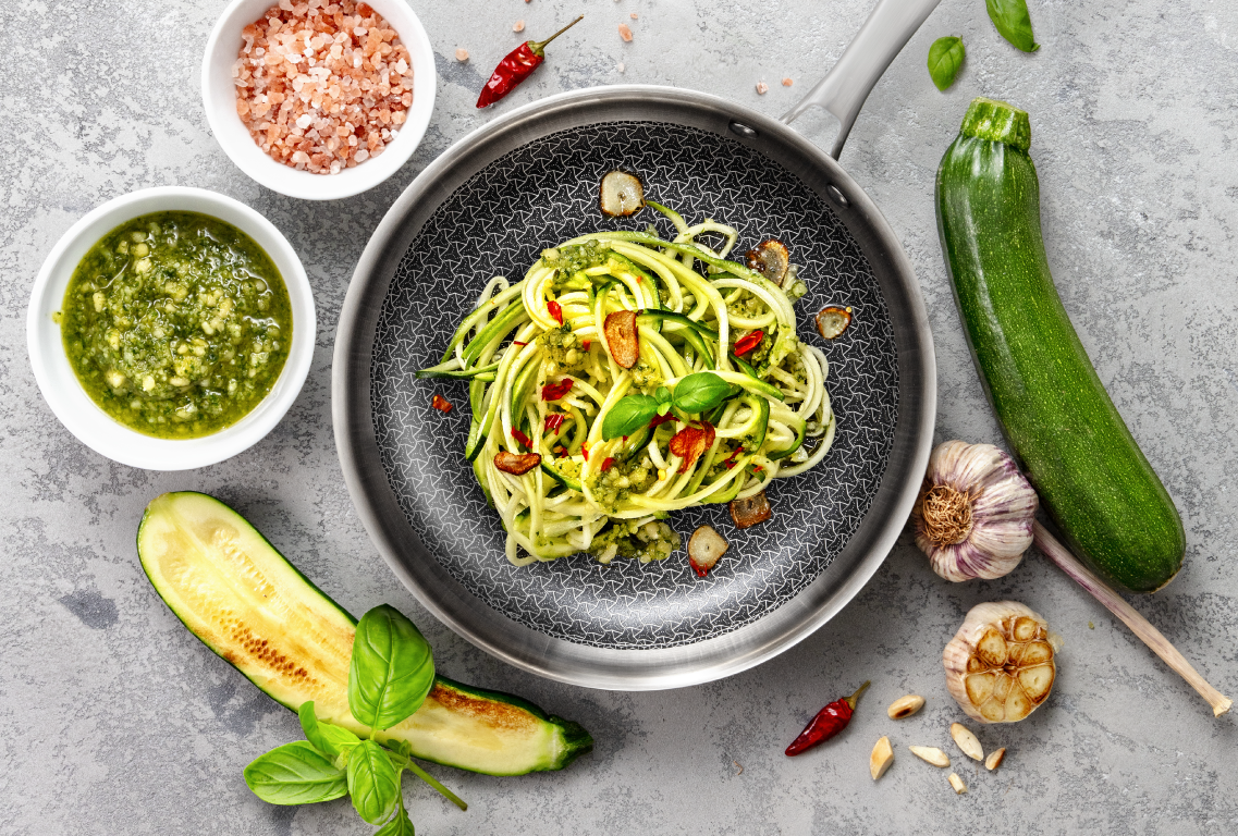 Obrazek przedstawia stalową patelnię z czarnym antyadhezyjnym dnem ze stalowym rastrem, na której leży spaghetti z cukinii z ziołami i listkami bazylii. Wokół patelni na szarym blacie leżą cukinie, główki czosnku, listki bazylii, pikantne papryczki, biała miseczka z solą himalajską i biała maseczka z zielonym pesto.