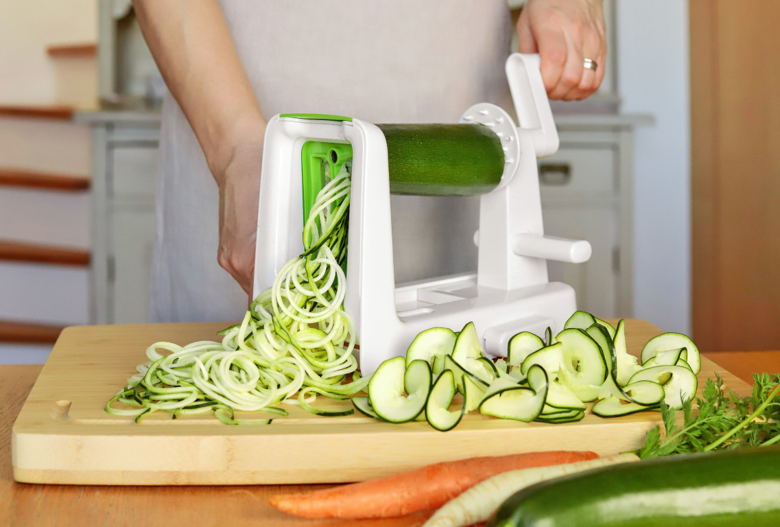 Ktoś robi spaghetti z cukinii w biało-zielonym spiralizerze. Obok leżą długie spirale przygotowane z cukinii.