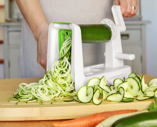 Ktoś robi spaghetti z cukinii w biało-zielonym spiralizerze. Obok leżą długie spirale przygotowane z cukinii.