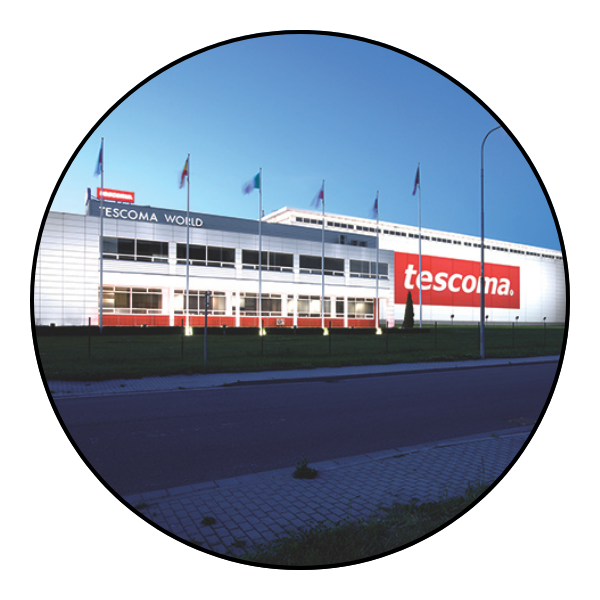 Zdjęcie siedziby Tescomy w Zlinie. Duży, niski, biały budynek, na którym widnieje duże czerwone logo tescoma i powiewają flagi różnych państw.