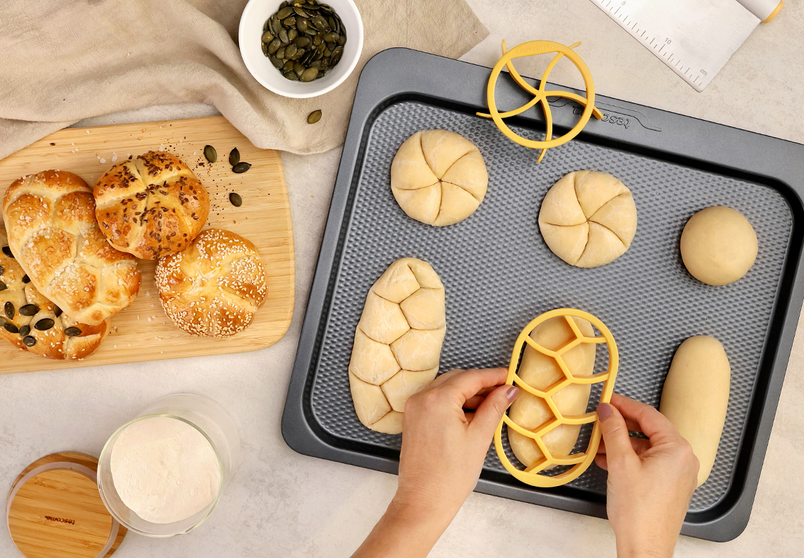 Żółte foremki do bułek DELÍCIA. Przy ich pomocy ktoś odbija wzór na surowych kajzerkach i chałkach, które leżą na płaskiej blasze do pieczenia. Po lewej od blachy, na drewnianej desce do krojenia leżą upieczone bułki posypane ziarnami.