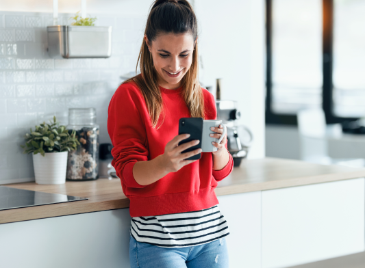 Kobieta stoi w kuchni, w jednej ręce trzyma kubek z napojem, a w drugiej smartfon, w który się wpatruje.