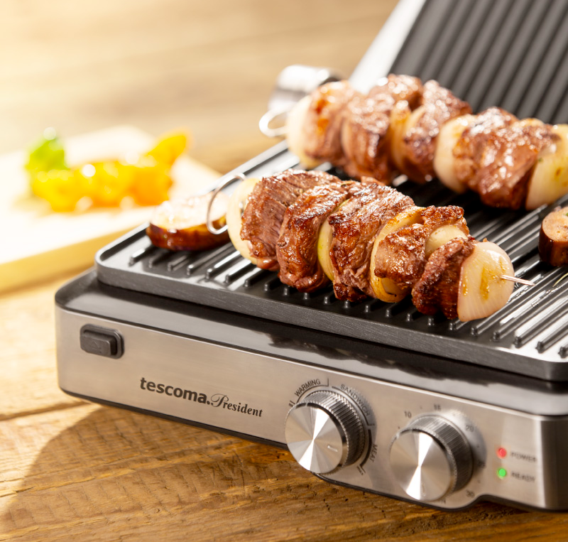 Opis: w otwartym srebrno-czarnym grillu kontaktowym leżą dwa szaszłyki z mięsem i cebulą. Grill ma dwa pokrętła, czerwoną i zieloną diodę oraz napis tescoma. President.