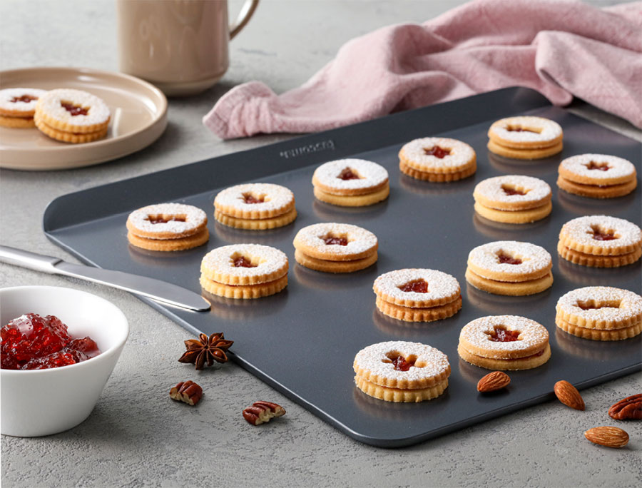 Plaska blacha do pieczenia, na której leżą kruche ciasteczka z nadzieniem z marmolady. Ciasteczka mają pośrodku otwory w świątecznych kształtach.